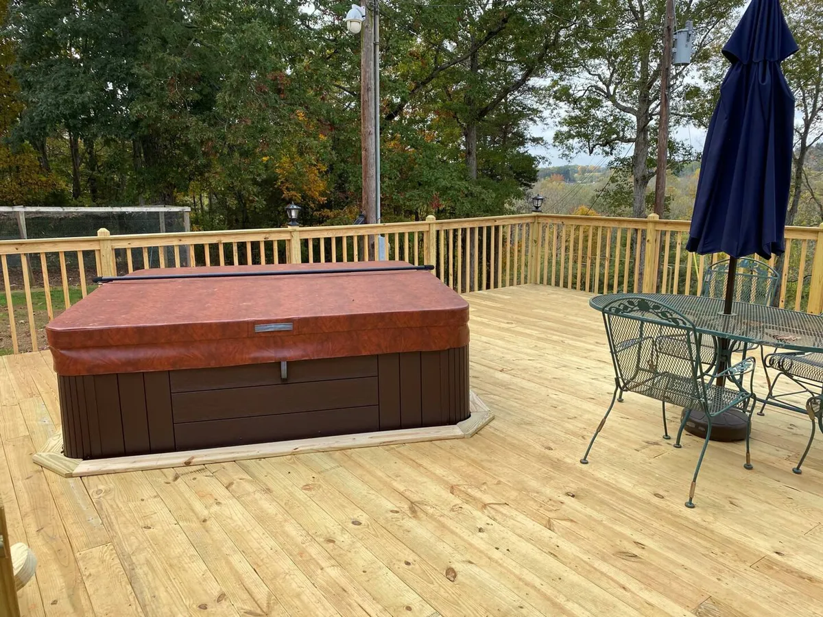 The image shows the exterior of a charming cottage nestled among tall trees, with a bubbling hot tub and an inviting outdoor dining set set on a spacious patio. The lush greenery creates a sense of seclusion and tranquility, while the setup suggests opportunities for stargazing, morning coffee, or evening gatherings.