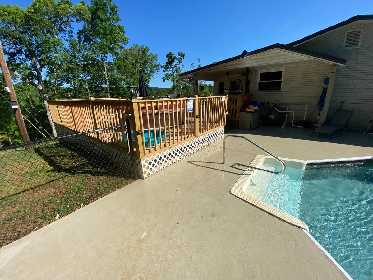 A broad view of the swimming pool flanked by a wooden deck, lush greenery, and the house exterior. The scene suggests leisure and social gatherings, with ample space for swimming, sunbathing, and outdoor activities, all set against a comfortable, home-like backdrop.