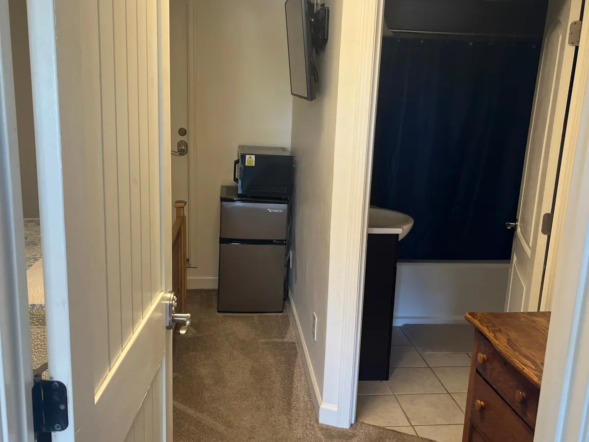 A practical and inviting living area showcasing a small refrigerator, a television, a wooden dresser, and a door leading to the rest of the cottage. The layout encourages relaxation, with convenient amenities at hand.