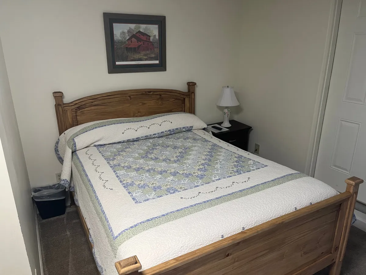 A warmly lit bedroom featuring a plush bed topped with a quilt, a softly glowing bedside lamp, and a tasteful painting on the wall. The overall ambiance is peaceful and welcoming, with natural light hinting at a restful atmosphere.