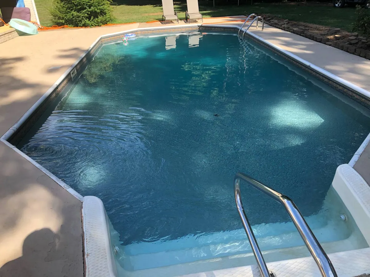 A tranquil outdoor swimming pool surrounded by lush greenery, featuring a stainless steel ladder for easy access. The pool area is inviting for swimming, relaxing, or sunbathing, and the peaceful ambiance is enhanced by the view of the natural landscape beyond the pool's edge.
