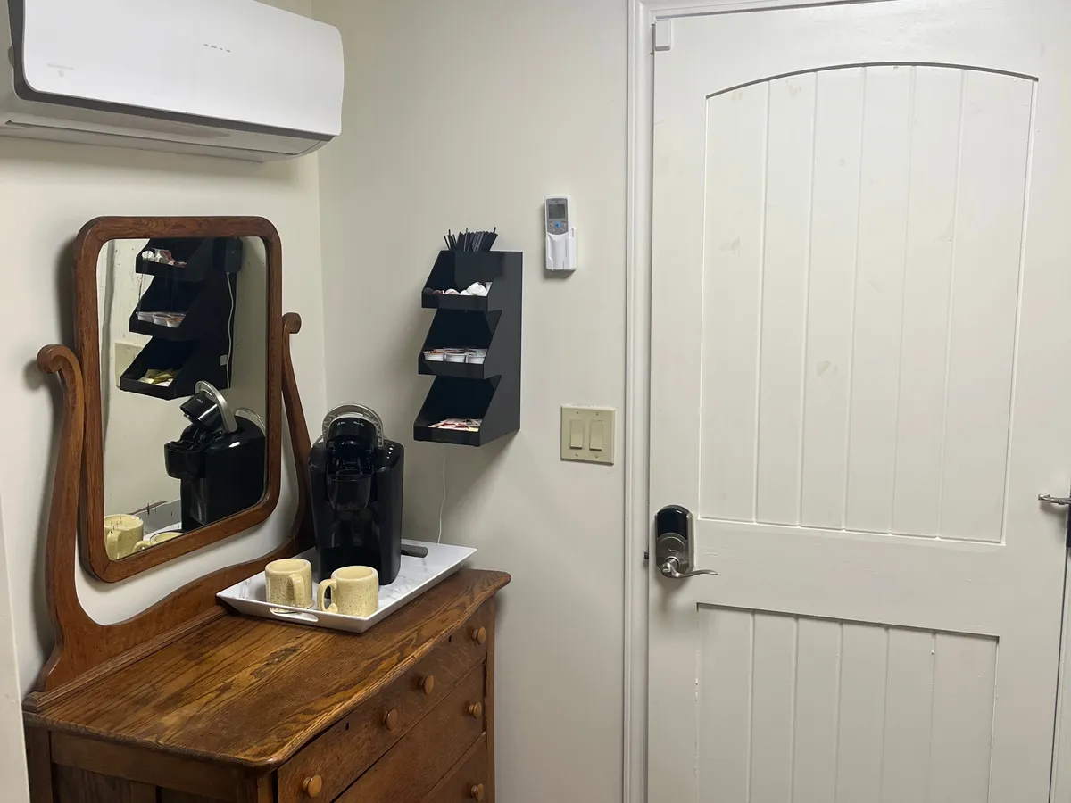 A bright, cozy interior scene featuring a wooden dresser crowned with a coffee machine, a large mirror reflecting soft natural light, and an accessory organizer. The nearby door hints at entryway access, while the neat arrangement creates a welcoming and lived-in atmosphere.