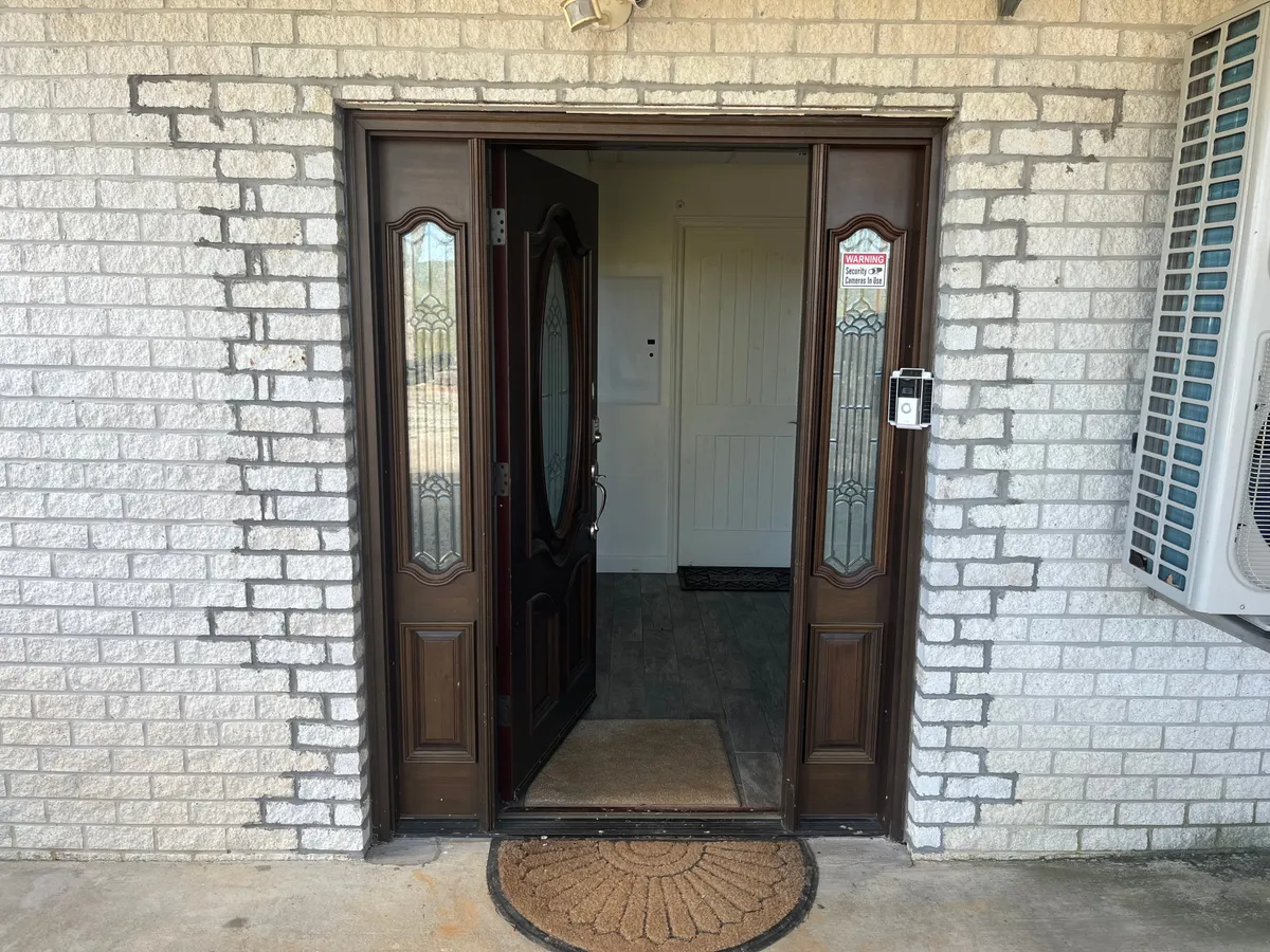 A welcoming entryway with double doors featuring decorative glass panels, set against a tidy exterior. A welcome mat and inviting design provide a sense of arrival and security within the gated property.
