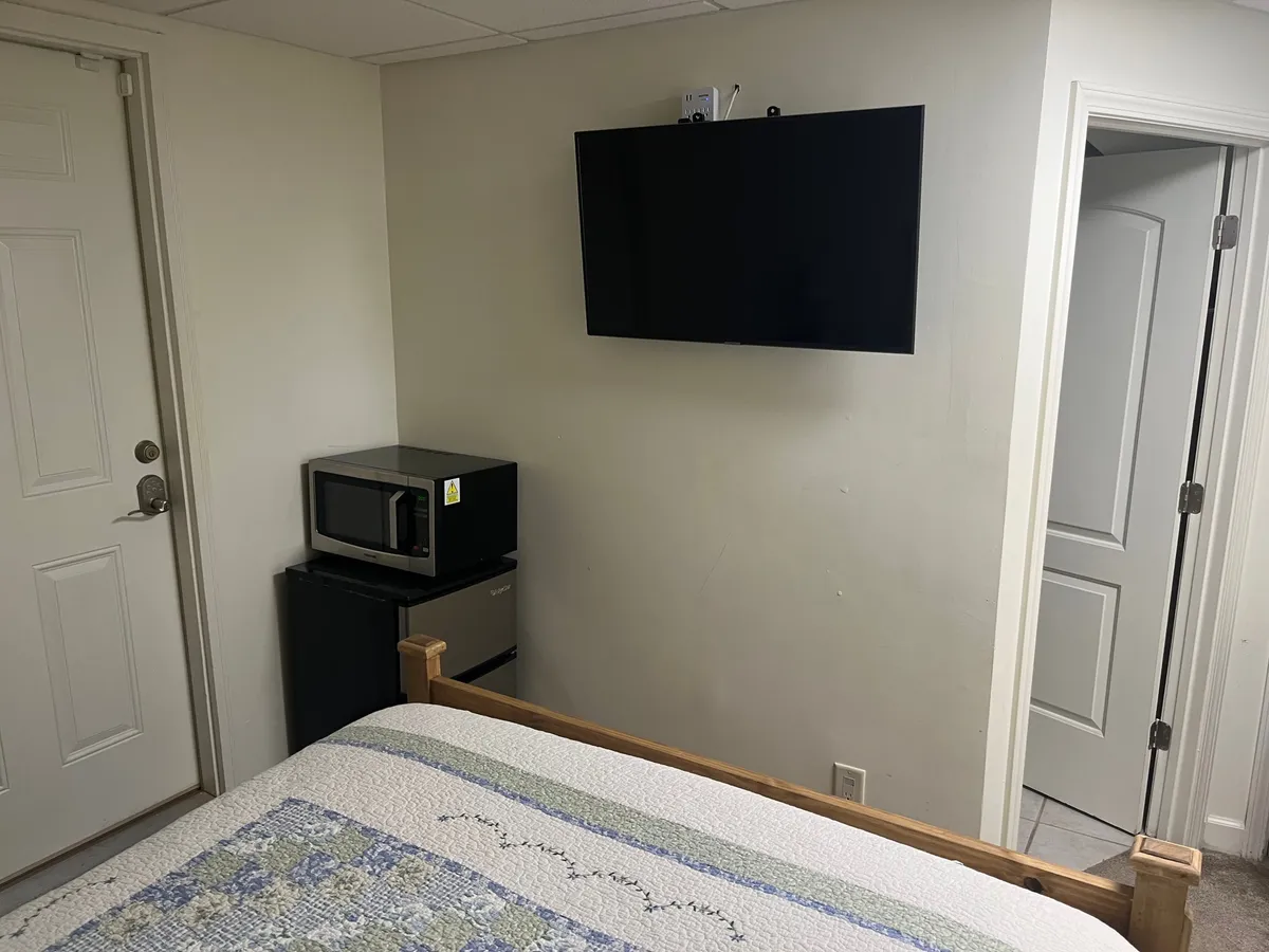 A comfortable bedroom space featuring a plush bed facing a television, with a microwave and small fridge nearby. A door at the edge of the frame suggests easy access and privacy, while the overall feeling is one of relaxation and convenience.